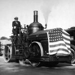 In 1929, the opening of this paved road was grand enough to warrant a celebration, as shown in the photograph. This patriotically decorated machine is a true steamroller (powered by a steam engine, at center)âand operated with Queens class by a Queens public works commissioner J. J. Halloran, who wears a three-piece suit and a tie.